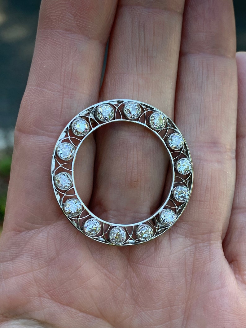 May include: A silver ring with a circular design featuring a row of round diamonds set in a delicate filigree pattern.