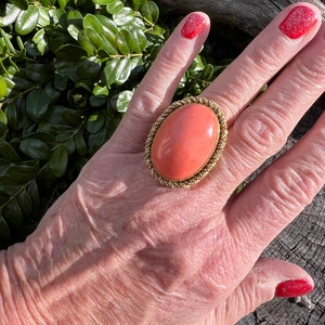 May include: A large oval ring with a coral-coloured stone set in a gold-toned metal frame. The ring is worn on a person's finger.