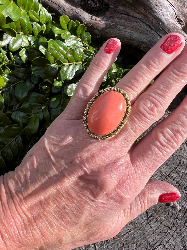 May include: A large oval ring with a coral-coloured stone set in a gold-toned metal frame. The ring is worn on a person's finger.