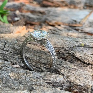 May include: A silver engagement ring with a large, round, clear diamond set in a prong setting. The band is decorated with a delicate leaf design.
