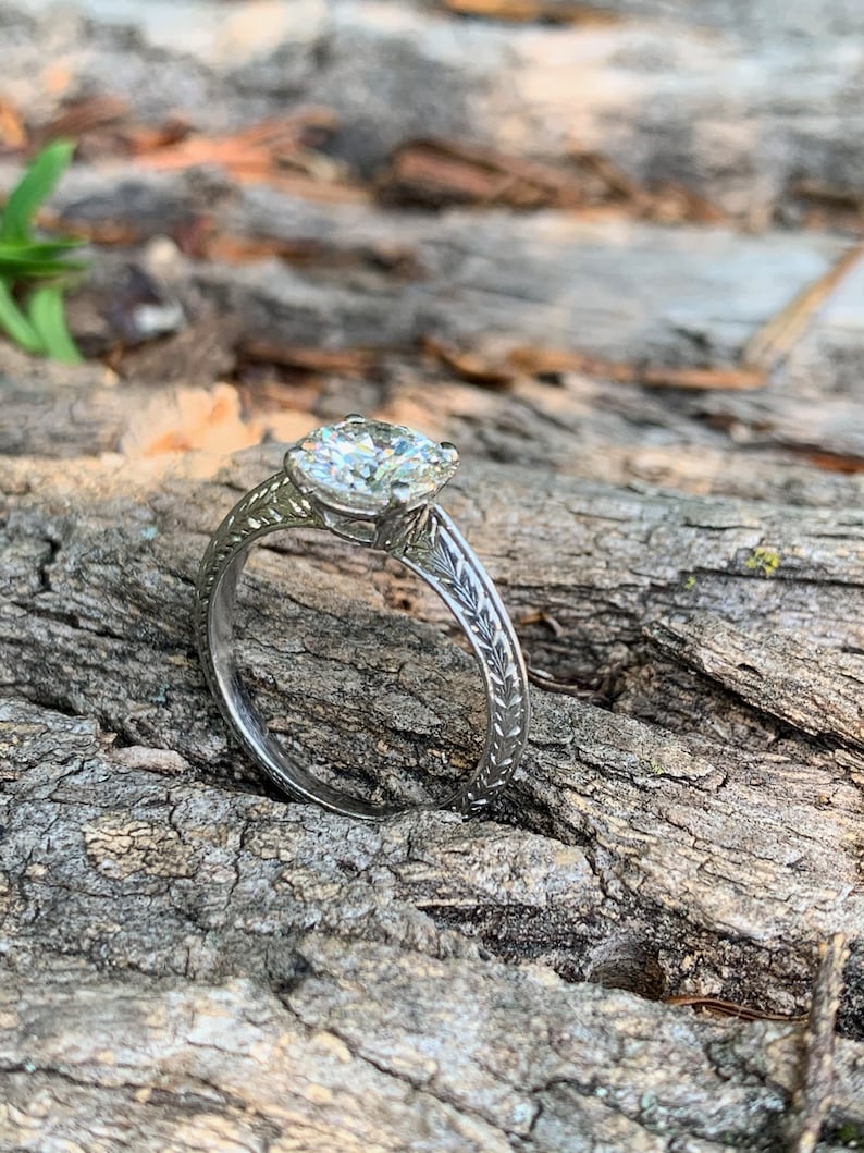 May include: A silver engagement ring with a large, round, clear diamond set in a prong setting. The band is decorated with a delicate leaf design.