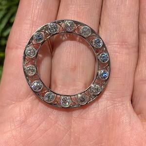 May include: A silver circular brooch with a delicate filigree design and a row of round, clear gemstones around the edge.
