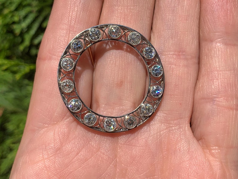 May include: A silver circular brooch with a delicate filigree design and a row of round, clear gemstones around the edge.