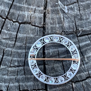 May include: A silver circular brooch with a delicate design and clear rhinestones around the edge. The brooch has a pin on the back for attaching to clothing.