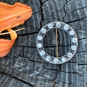 May include: A circular brooch with a silver setting and a row of clear round stones around the edge. The brooch has a gold pin.