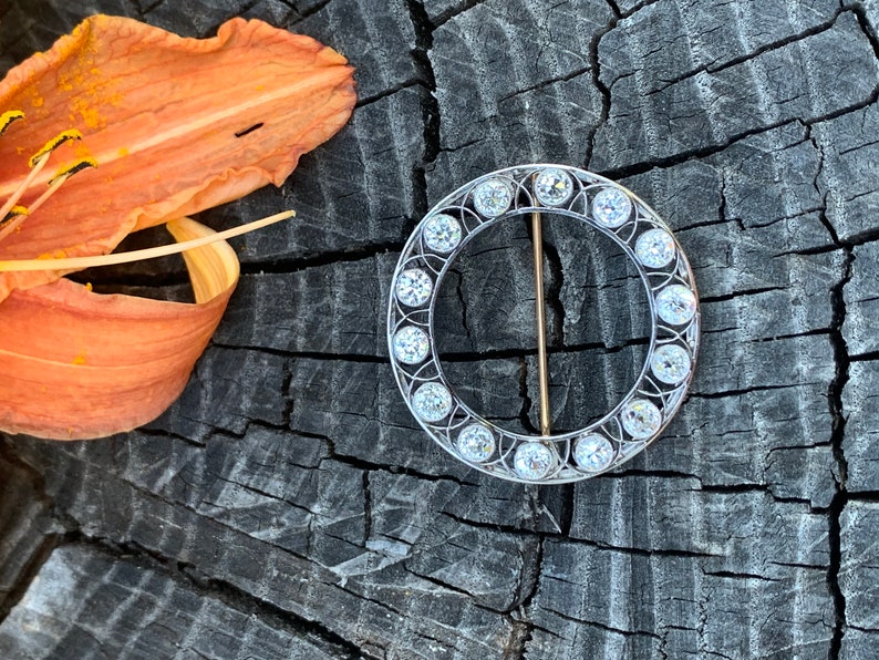 May include: A circular brooch with a silver setting and a row of clear round stones around the edge. The brooch has a gold pin.