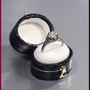 May include: A silver ring with a round, clear stone set in a floral design, displayed in a black ring box with a white interior.