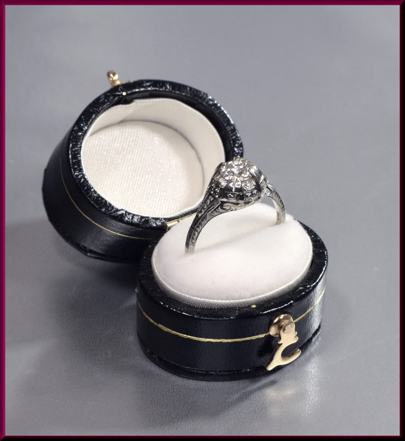 May include: A silver ring with a round, clear stone set in a floral design, displayed in a black ring box with a white interior.