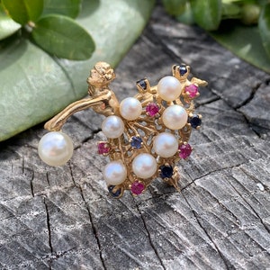 May include: A gold-toned brooch featuring a stylized figure with a pearl-encrusted skirt and a scattering of red and blue gemstones.