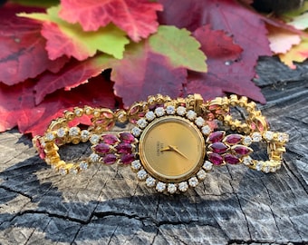 May include: A gold wristwatch with a round face and a diamond-encrusted band. The watch face is gold with black hands and the word "Concord" printed in black. The band is adorned with small diamonds and red rubies.