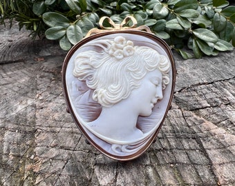 May include: A cameo brooch featuring a carved portrait of a woman with a floral hairpiece. The cameo is set in a gold tone metal frame with a bow detail.