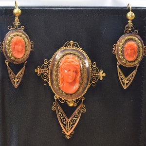 May include: A set of antique coral cameo jewelry featuring a gold filigree design. The set includes a brooch and a pair of earrings. Each piece features a carved coral cameo of a woman's face.