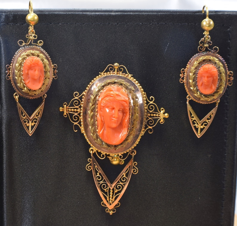 May include: A set of antique coral cameo jewelry featuring a gold filigree design. The set includes a brooch and a pair of earrings. Each piece features a carved coral cameo of a woman's face.