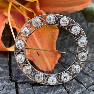 May include: A silver circular brooch with a delicate filigree design and a row of round, clear gemstones around the edge.