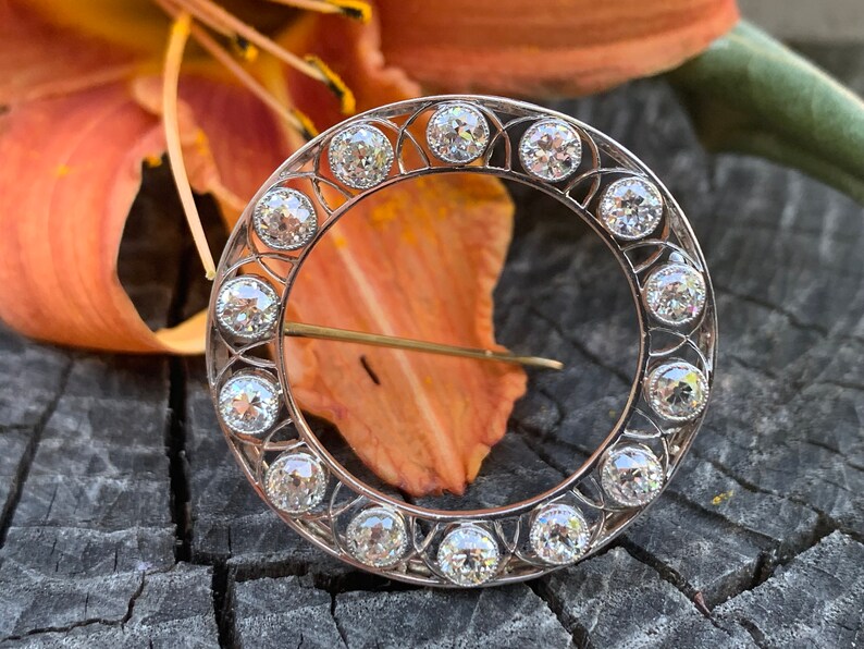 May include: A silver circular brooch with a delicate filigree design and a row of round, clear gemstones around the edge.