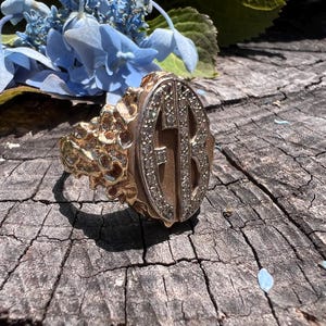 Puede incluir: Un anillo de oro con un gran engaste ovalado que presenta un diseño de monograma con diamantes. El anillo tiene una banda texturizada.