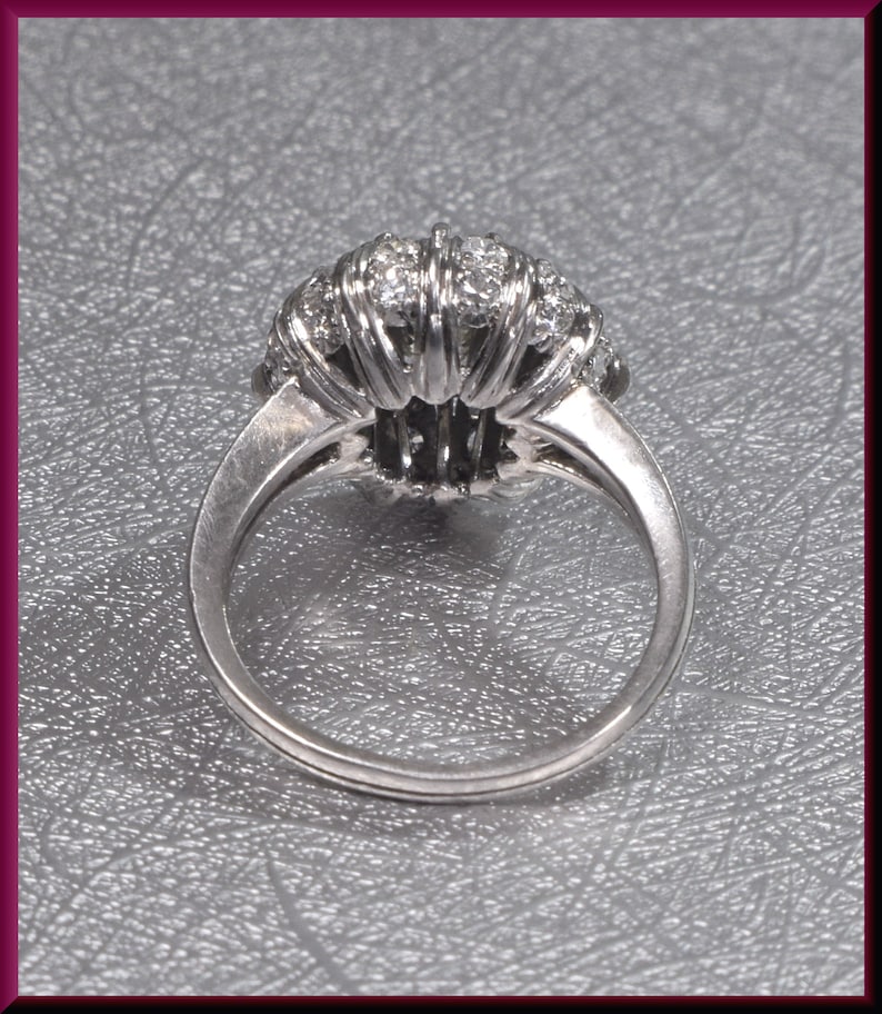May include: A silver ring with a large, round, diamond-encrusted setting. The ring is set on a gray textured surface.