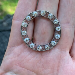 May include: A silver circular brooch with a delicate filigree design and a ring of round, clear gemstones.