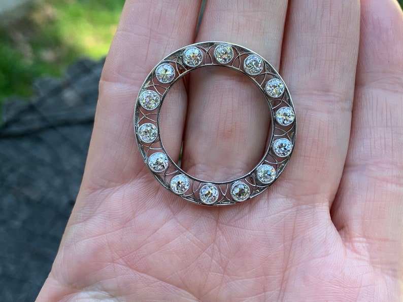 May include: A silver circular brooch with a delicate filigree design and a ring of round, clear gemstones.