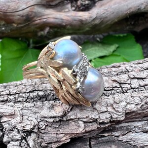 Könnte beinhalten: Ein goldener Ring mit zwei großen grauen Perlen und kleinen Diamanten, die in einem Blattdesign eingefasst sind.