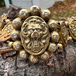 Op de afbeelding: Een gouden armband met een leeuwenkopbedel en meerdere ronde bedels met heldere stenen. De armband ligt op een houten oppervlak.