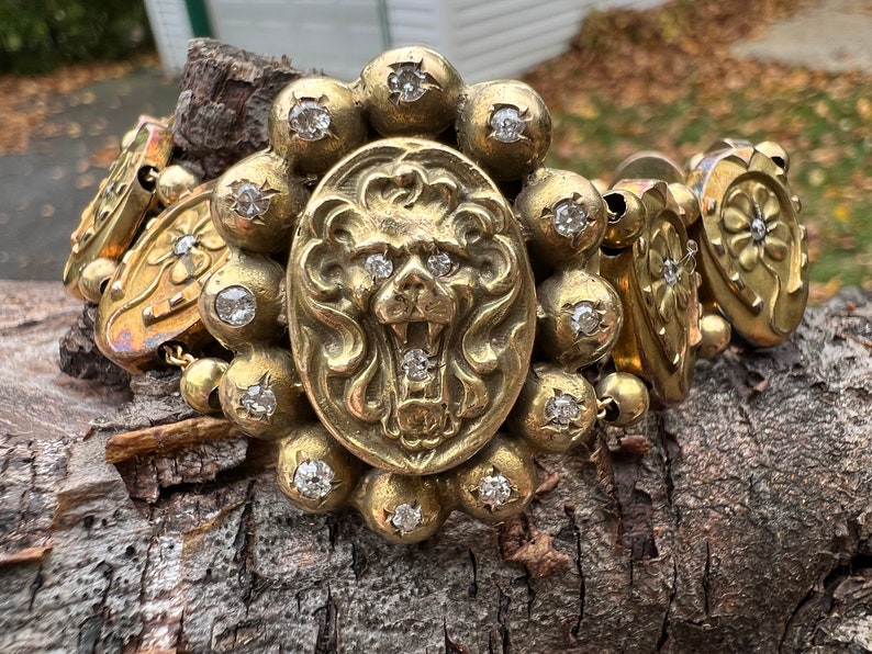 Op de afbeelding: Een gouden armband met een leeuwenkopbedel en meerdere ronde bedels met heldere stenen. De armband ligt op een houten oppervlak.