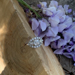 May include: A silver ring with a large round diamond surrounded by smaller diamonds. The ring is set on a piece of wood with purple flowers.