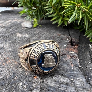 May include: A gold-toned Penn State University class ring with a blue center stone. The ring features the words "PENN STATE" and a Nittany Lion emblem. The ring is set against a rustic wooden surface with green foliage.