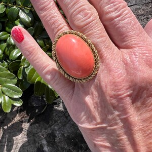 May include: A large oval coral ring with a gold setting. The ring is on a person's finger.