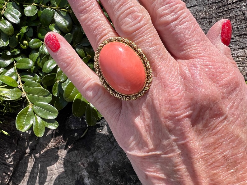 May include: A large oval coral ring with a gold setting. The ring is on a person's finger.