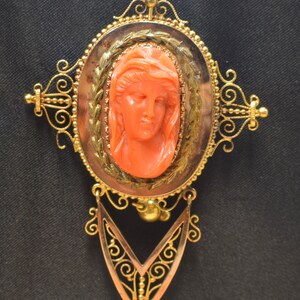 May include: A gold filigree brooch with a coral cameo of a woman's face. The cameo is set in a gold oval frame with a detailed floral design.