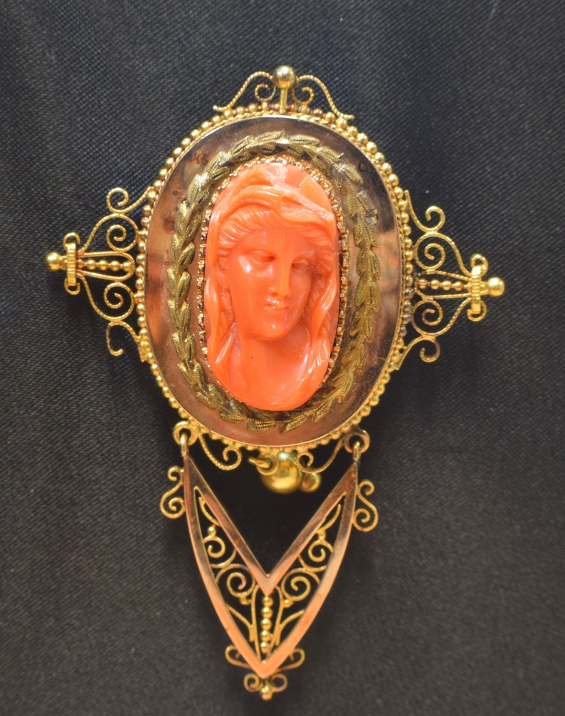 May include: A gold filigree brooch with a coral cameo of a woman's face. The cameo is set in a gold oval frame with a detailed floral design.