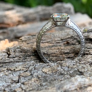 May include: A silver engagement ring with a large, clear, round diamond set in a prong setting. The band is decorated with a delicate, engraved floral design.
