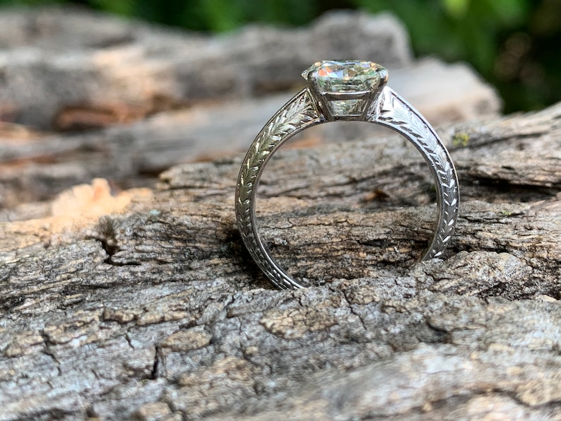 May include: A silver engagement ring with a large, clear, round diamond set in a prong setting. The band is decorated with a delicate, engraved floral design.