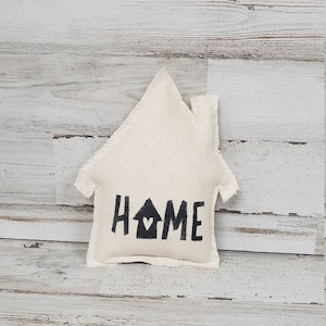 May include: A white fabric house-shaped pillow with the word "HOME" in black lettering and a small heart in the center of the "O".