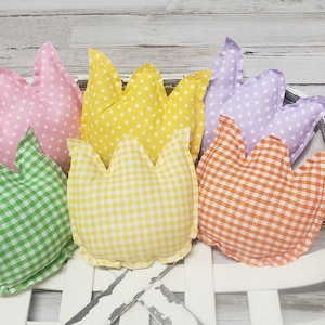 May include: A collection of colorful, tulip-shaped fabric decorations in a white distressed basket. The tulips feature various patterns, including polka dots and gingham, in shades of pink, yellow, green, orange, and purple. The basket sits on a white wooden surface.