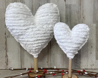 May include: Two white, plush heart decorations on wooden stands. The hearts have a textured, fluffy appearance and are mounted on natural wood slices. Decorative branches with red and white berries are in the foreground.