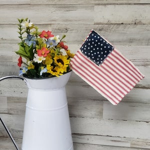 May include: A white pitcher with a black rim filled with a bouquet of red, white, and blue flowers. A small American flag with a blue star field is hanging from the pitcher's handle.