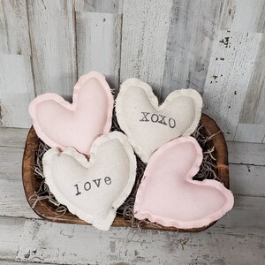 May include: Four heart-shaped fabric ornaments in a wooden bowl. Two are light pink, and two are off-white. The off-white hearts have the words "love" and "XOXO" printed on them in black. The bowl is filled with dried moss.