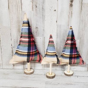 Puede incluir: Tres árboles de Navidad decorativos de diferentes tamaños. Cada árbol presenta un diseño de tela a cuadros en rojo, azul, verde y amarillo. Los árboles están montados sobre clavijas de madera y bases circulares de madera, creando una estética rústica.