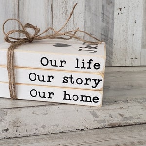 May include: Three white wooden blocks stacked on top of each other, tied together with twine. The blocks have black lettering that reads "Our life", "Our story", and "Our home".
