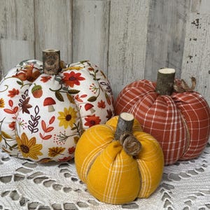 Puede incluir: Tres calabazas de tela de diferentes tamaños. La más grande es blanca con un estampado floral de girasoles amarillos, hojas naranjas y setas rojas. Las otras dos son de cuadros naranjas y rojos. Cada una tiene un tallo de madera.