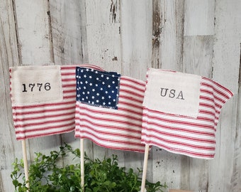 May include: Three small American flag-themed fabric pennants on sticks. The pennants are red, white, and blue with the numbers "1776" on one, "USA" on another, and a small blue star field on the third.