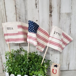 May include: Three small American flag bunting with red and white stripes and blue stars. The flags are on wooden sticks and are in a white planter with green plants. The flags have the words "USA", "1776" and a blue star on them.
