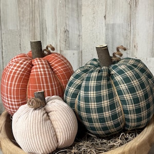 Puede incluir: Tres calabazas de tela en un cuenco de madera. Las calabazas son de cuadros naranjas, cuadros verdes y pana crema. Cada calabaza tiene un tallo de madera y detalles de cuerda.