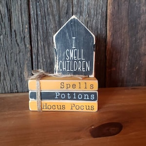 Op de afbeelding: Een houten Halloween-decoratie met een zwart huisvormig bord met de tekst "I SMELL CHILDREN". Daaronder staan drie gestapelde blokken met de tekst "Spells", "Potions" en "Hocus Pocus", samengebonden met touw. De blokken zijn oranje en zwart.
