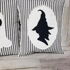 May include: Two black and white striped pillow covers with a white oval silhouette of a witch on one pillow. The pillows are on a whitewashed wood surface.