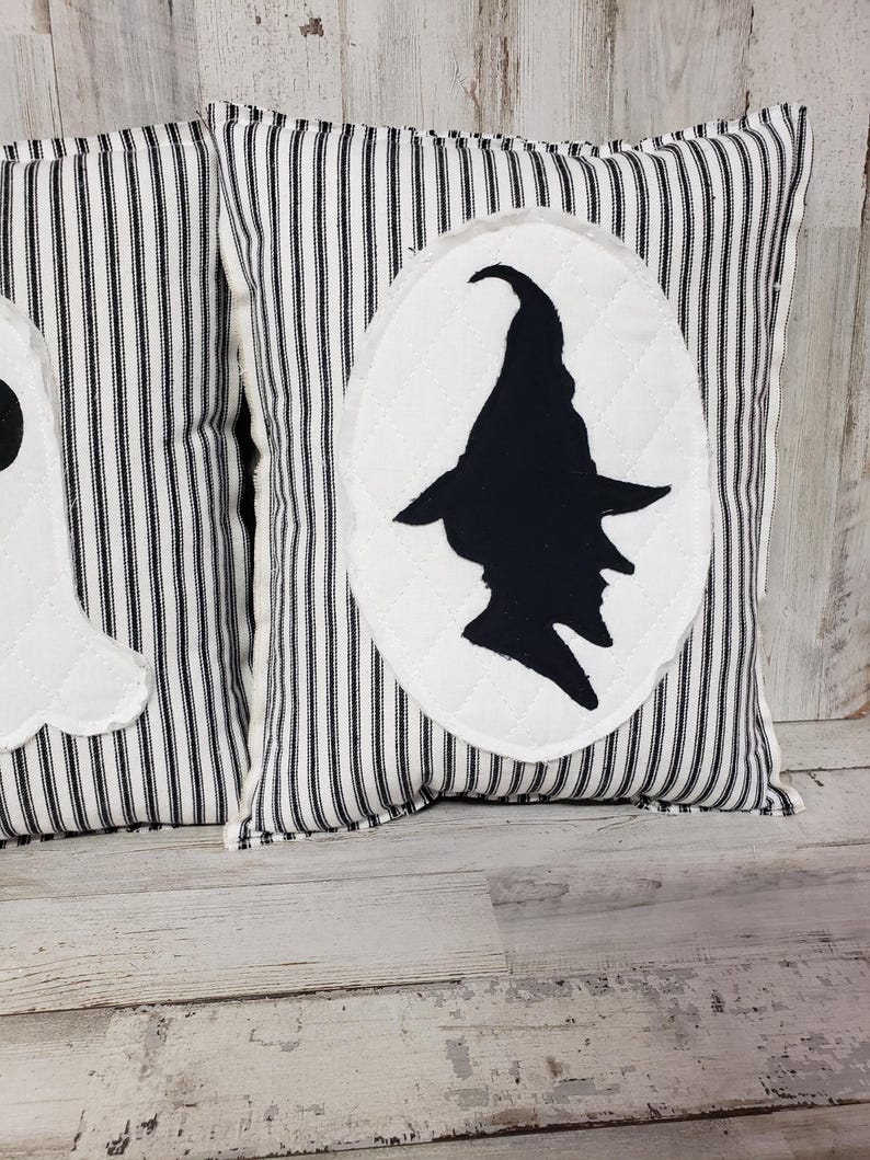 May include: Two black and white striped pillow covers with a white oval silhouette of a witch on one pillow. The pillows are on a whitewashed wood surface.
