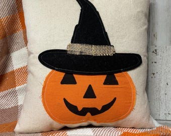 May include: A square decorative pillow with a Halloween theme. The pillow features an orange pumpkin face with a black witch hat. The pillow is on a plaid blanket with orange, white, and brown colors.