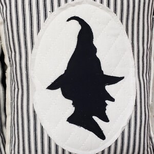 May include: Black and white striped pillow with a white oval patch featuring a black silhouette of a witch wearing a pointed hat.